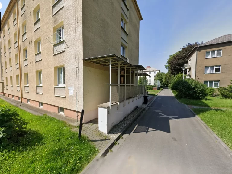 Úzká ulice lemovaná stromy vedle béžového vícepatrového činžovního domu s malou oplocenou terasou. Vlevo je travnatá plocha a v pozadí pod jasně modrou oblohou je vidět další obytná budova.