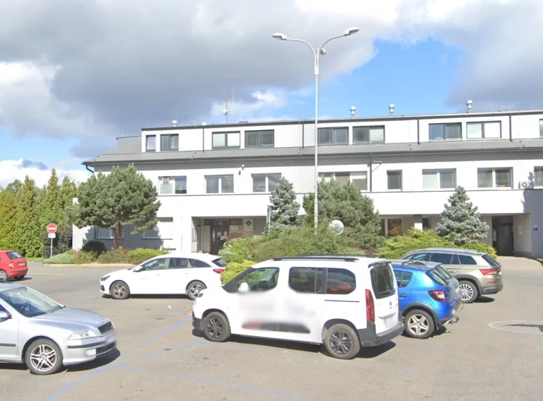 Parkoviště s několika auty před dvoupatrovou budovou s bílými zdmi a velkými okny. Budova je obklopena zelenými keři a stromy pod polojasnou oblohou.