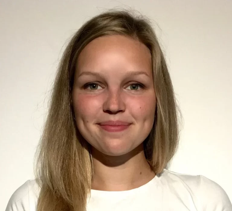 Žena s dlouhými blond vlasy a dolíčky se lehce usmívá, má na sobě bílou košili na světlém pozadí.