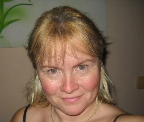 Osoba s blond vlasy a lehkým úsměvem je zobrazena na detailním portrétu. Na pozadí je částečně viditelné zelené a bílé umělecké dílo.
