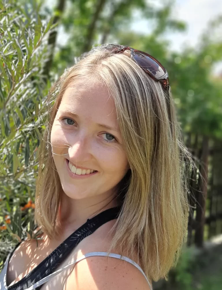 Člověk s blond vlasy, nosí sluneční brýle na hlavě, se venku usmívá. Jsou blízko nějakých zelených rostlin a oranžových květů, přičemž jejich tvář ozařuje sluneční světlo. Pozadí je rozmazané a zaměřuje se na osobu.